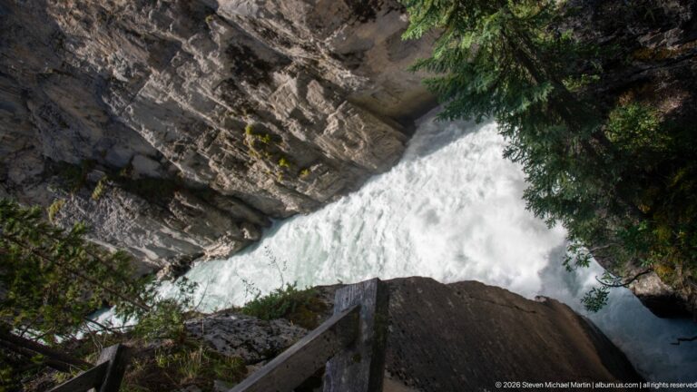 Downward view past a wooden railing to foaming water beneath layered rock walls and conifer branches.