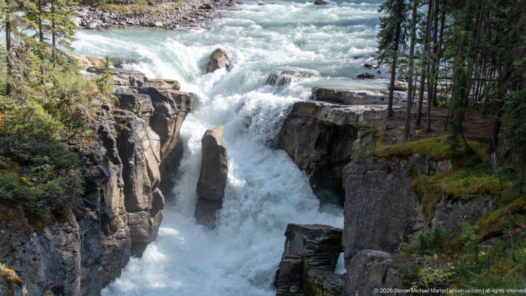 Broad view of white water descending around a central rock between steep canyon walls and conifers.