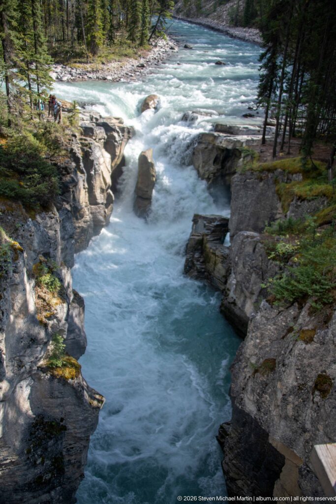 Turquoise river narrows into a white-water drop around a central rock, enclosed by forested rock walls.