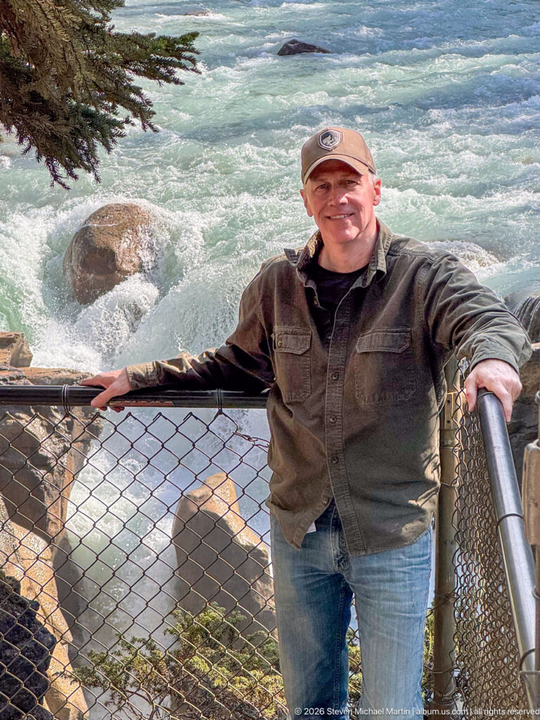 Portrait of a visitor at a fenced overlook with turbulent turquoise river water and rocks behind him.