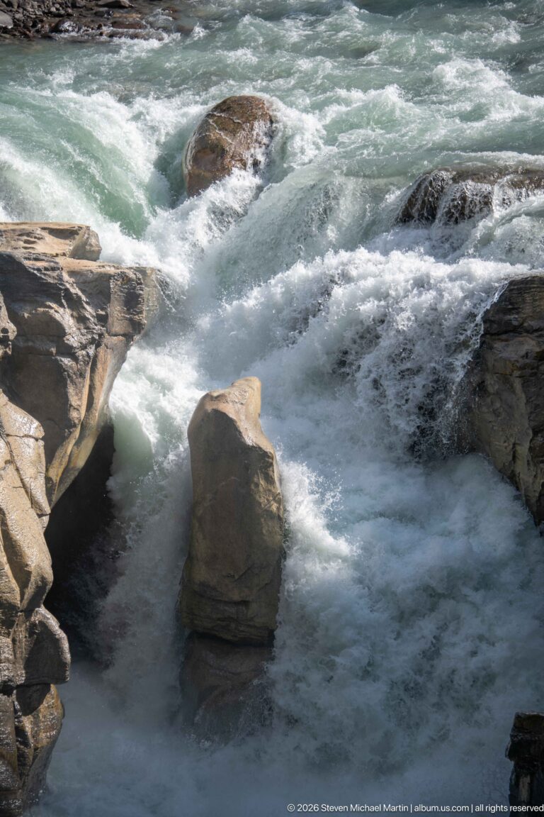 Close view of blue-green rapids breaking around a tall angular rock and exposed bedrock.