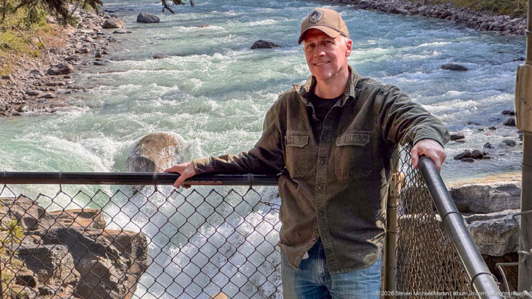 Full-length visitor portrait at a fenced overlook above fast glacial water and a rocky channel.