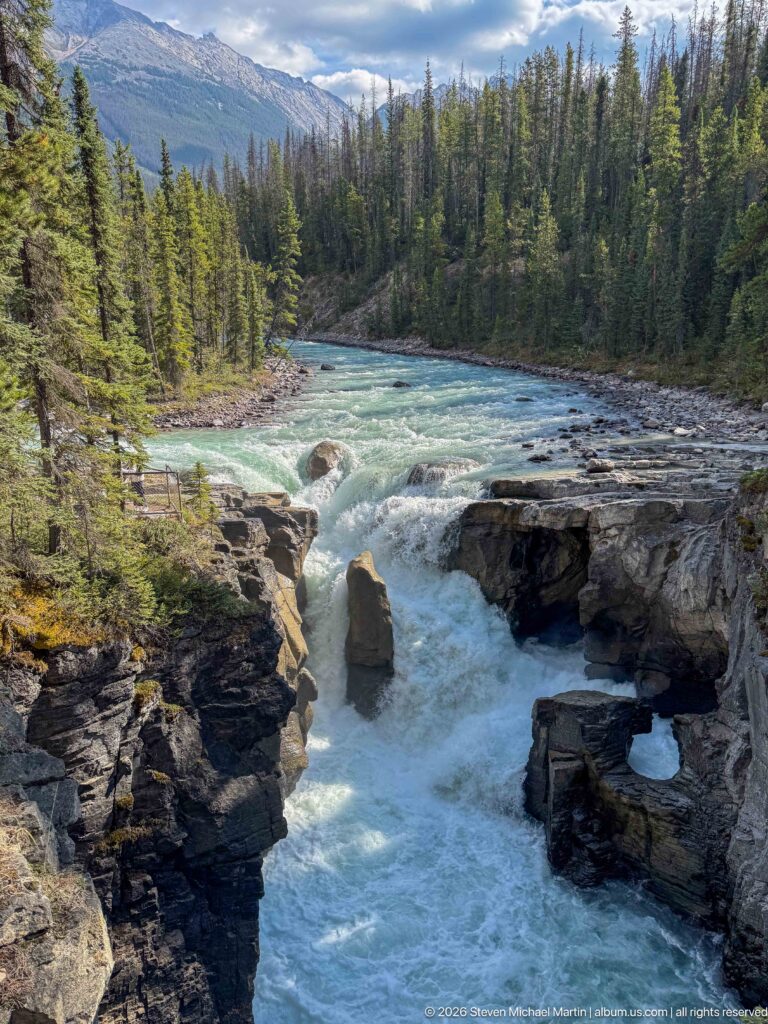 Turquoise river plunging through a narrow rocky gorge surrounded by evergreen forest and mountains.