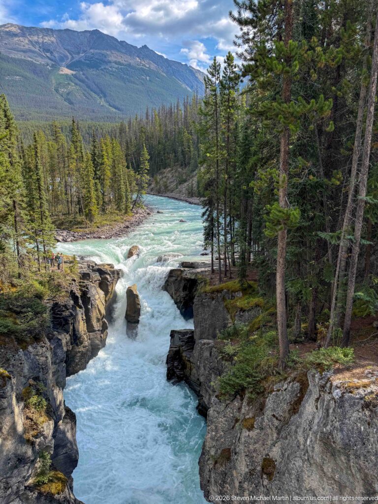 Elevated view of turquoise water dropping into a narrow rocky gorge amid conifers and mountain slopes.
