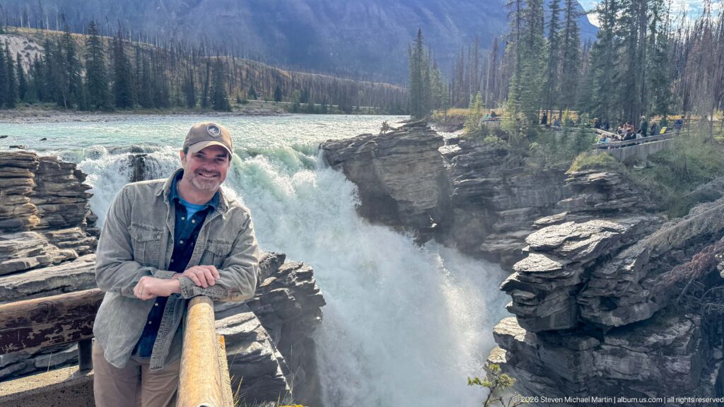 Man in a cap leaning on a wooden railing with Athabasca Falls and a crowded viewpoint behind him.