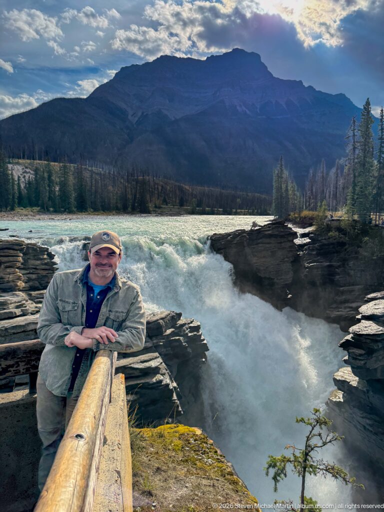 Man leaning on a wooden railing with Athabasca Falls, turquoise river, forest, and mountains behind him.