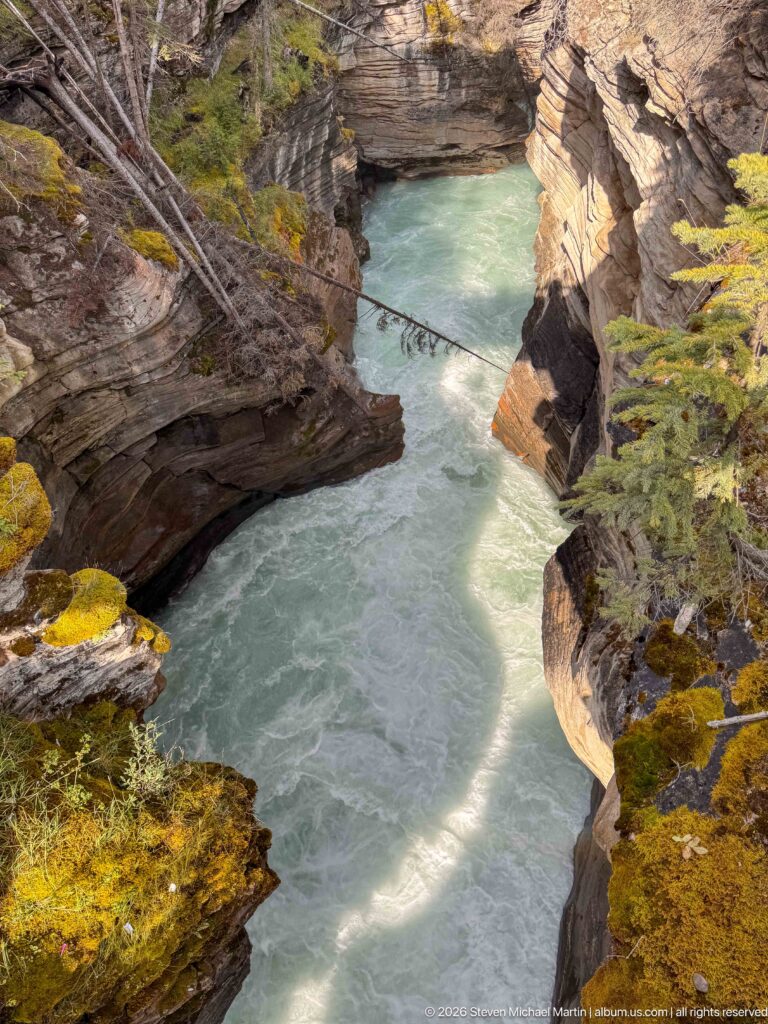 Turquoise river pool enclosed by steep sculpted bedrock, with mossy canyon edges and a band of sunlight.