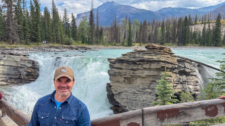 Smiling man in cap at a railing with Athabasca Falls, layered rock, forest, and mountains behind him.