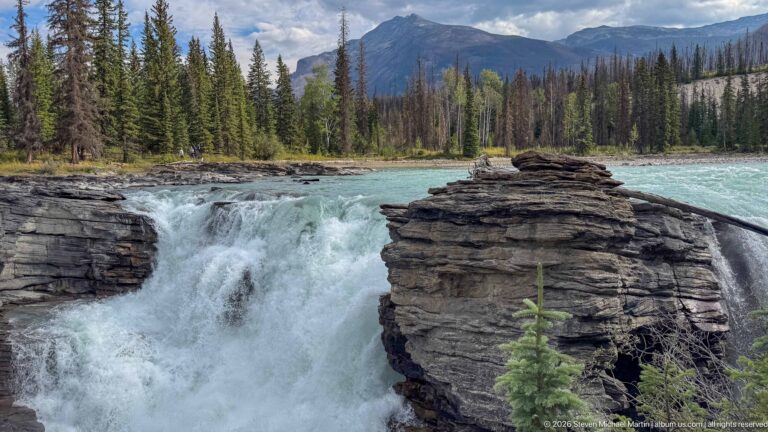 Close view of Athabasca Falls rushing around a tall layered rock outcrop, with turquoise river and forest beyond.