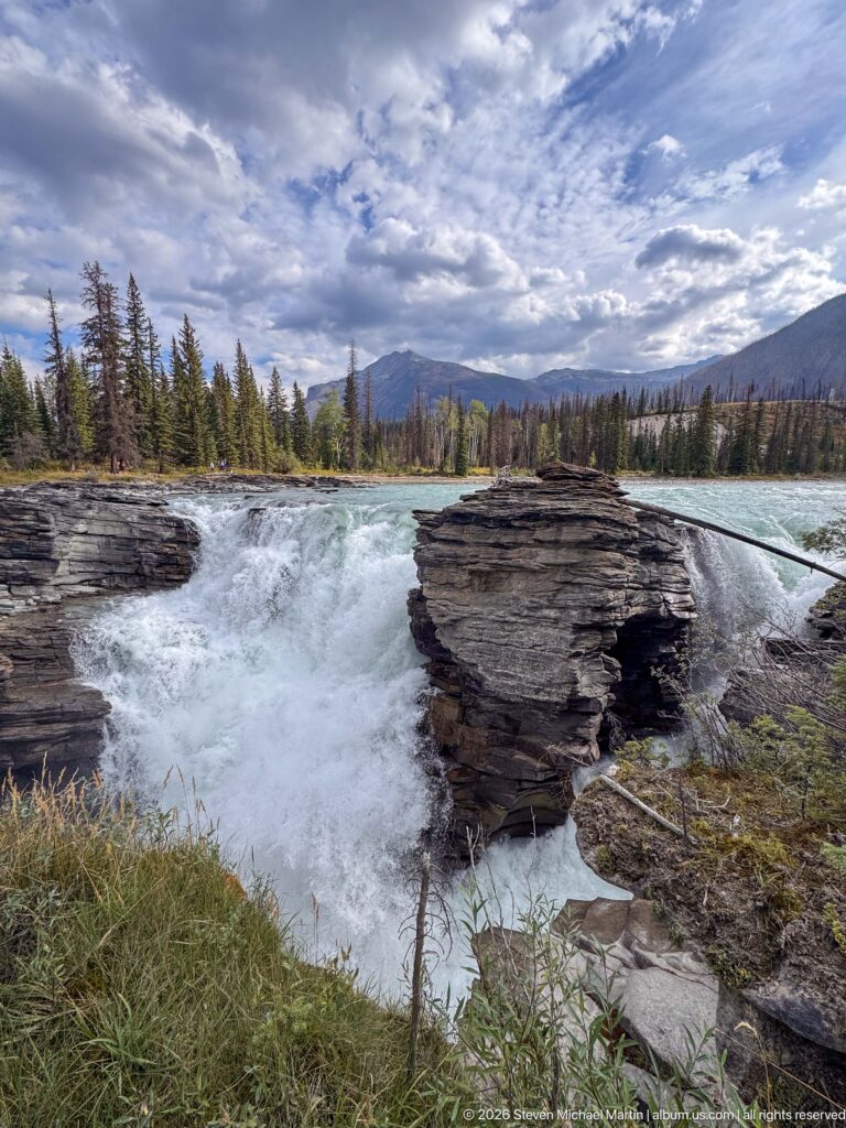 Wide view of whitewater between rock shelves with grass, conifer forest, distant mountains, and dramatic clouds.