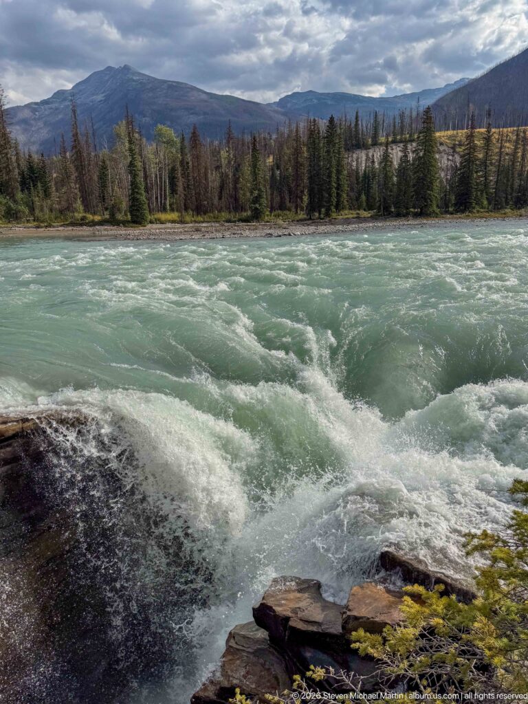 Turquoise rapids filling a broad river channel with a rocky foreground, conifer forest, and mountains beyond.