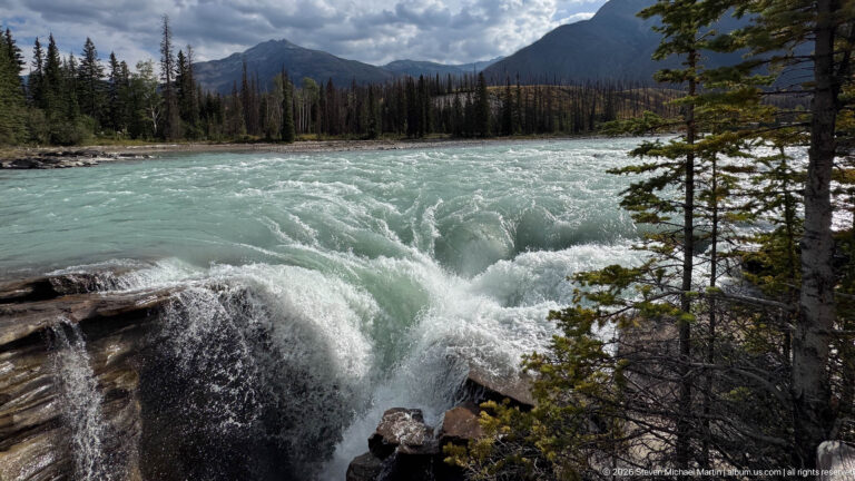 Turquoise river rapids beside a rocky ledge and conifers, with forest and a mountain slope in the distance.
