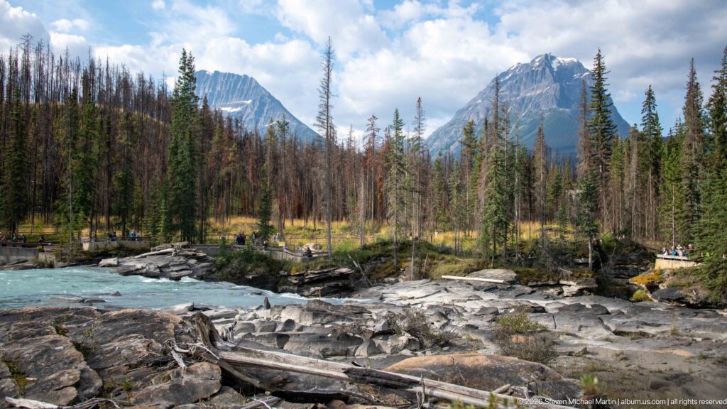 Turquoise river beside broad bedrock shelves and fallen logs, with conifer forest and steep mountains beyond.