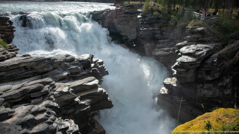 Powerful white waterfall plunging into a narrow layered-rock gorge, with visitors on the far rim.