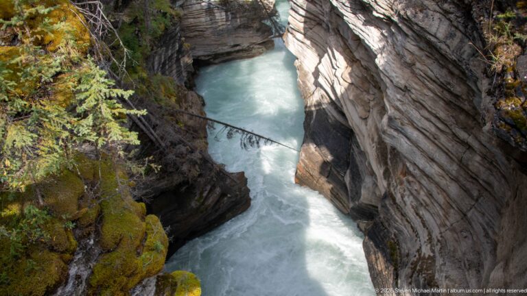 Downward view of pale turquoise river squeezed between steep layered canyon walls with moss along the rim.