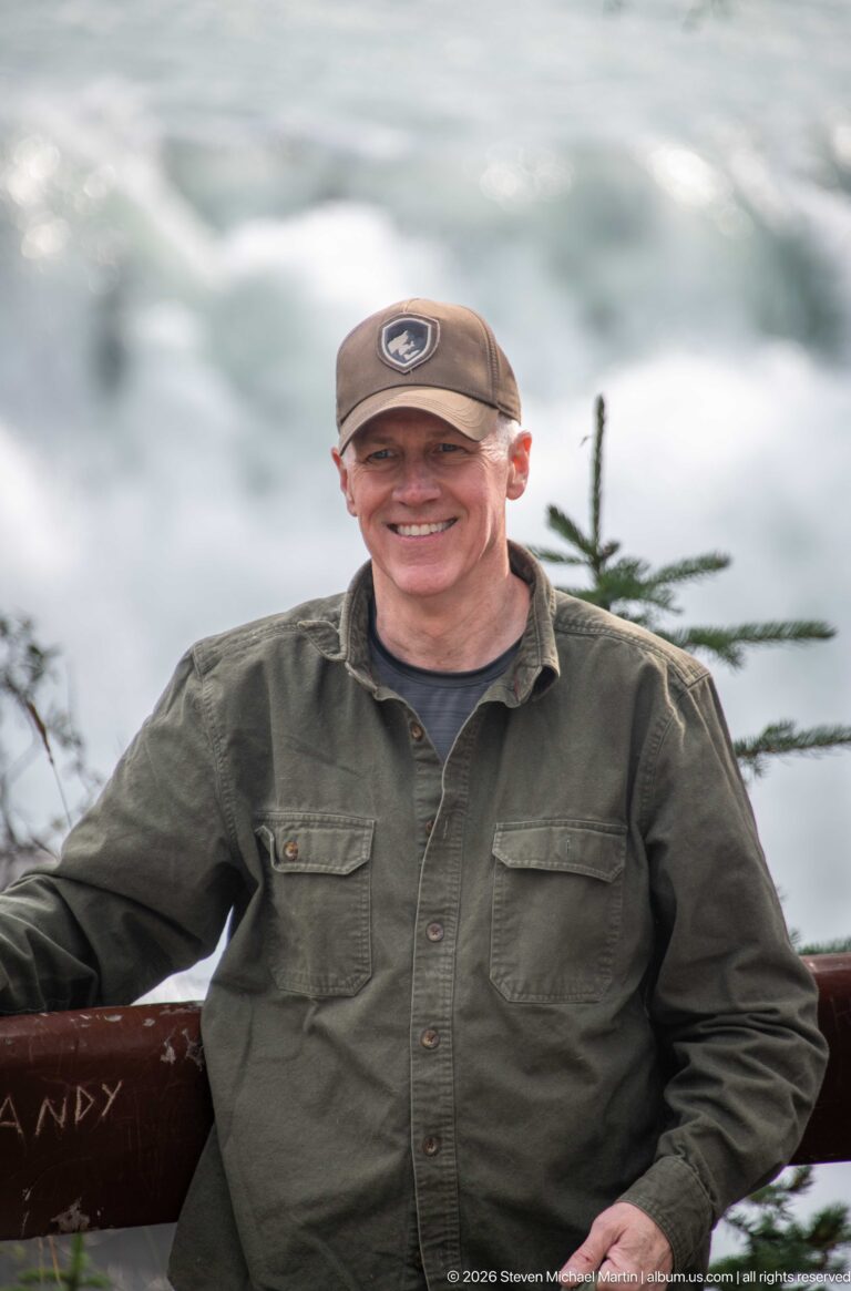 Smiling man in a cap and green shirt beside a wooden railing, with blurred whitewater behind him.
