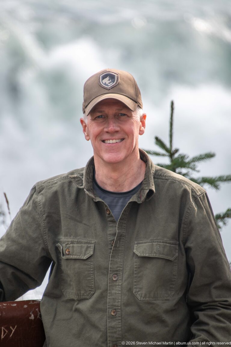 Close portrait of a smiling man in a cap and glasses, with blurred river water and a conifer behind.