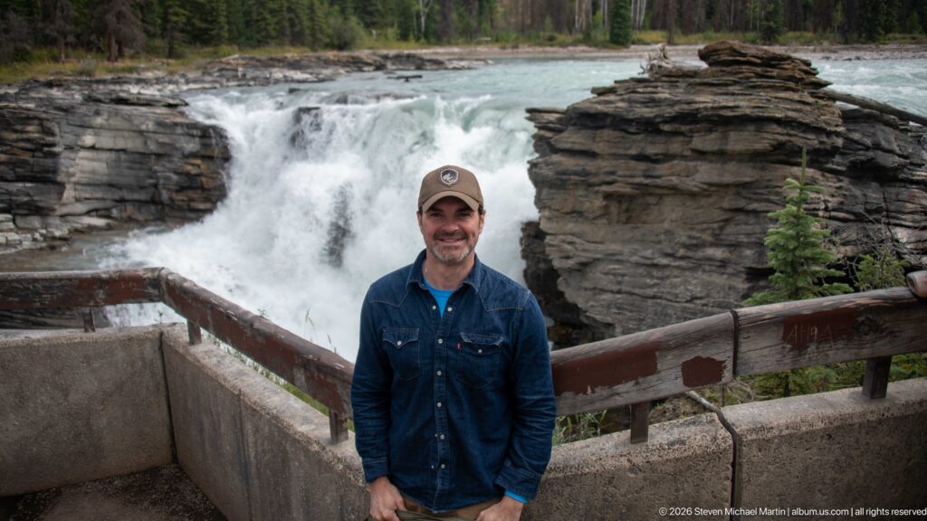 Smiling man in cap and blue shirt at a railing, with Athabasca Falls and layered rock directly behind.