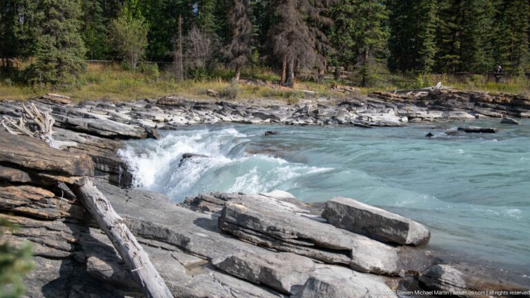 Turquoise river rapids curling around layered rock slabs and a large block, with driftwood and forest beyond.