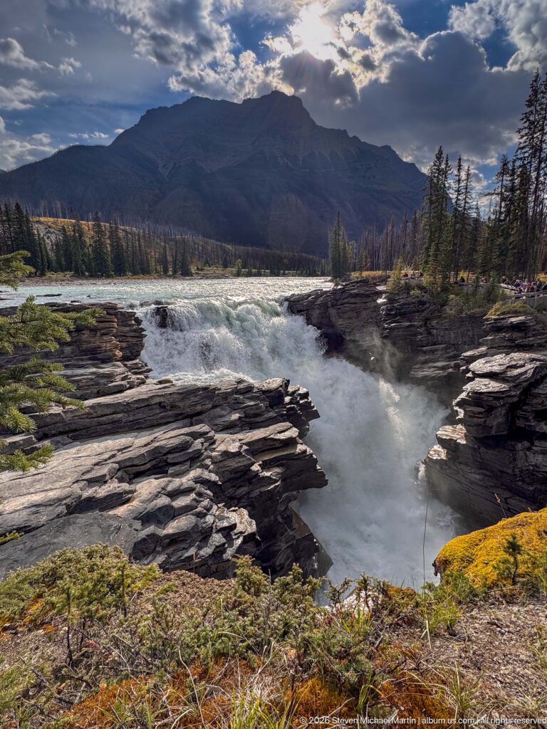 Wide view of turquoise river water plunging between layered rock shelves, with forest and mountains beyond.