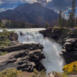 Powerful waterfall plunging into a narrow rocky gorge beneath a mountain, with forest and visitors at right.