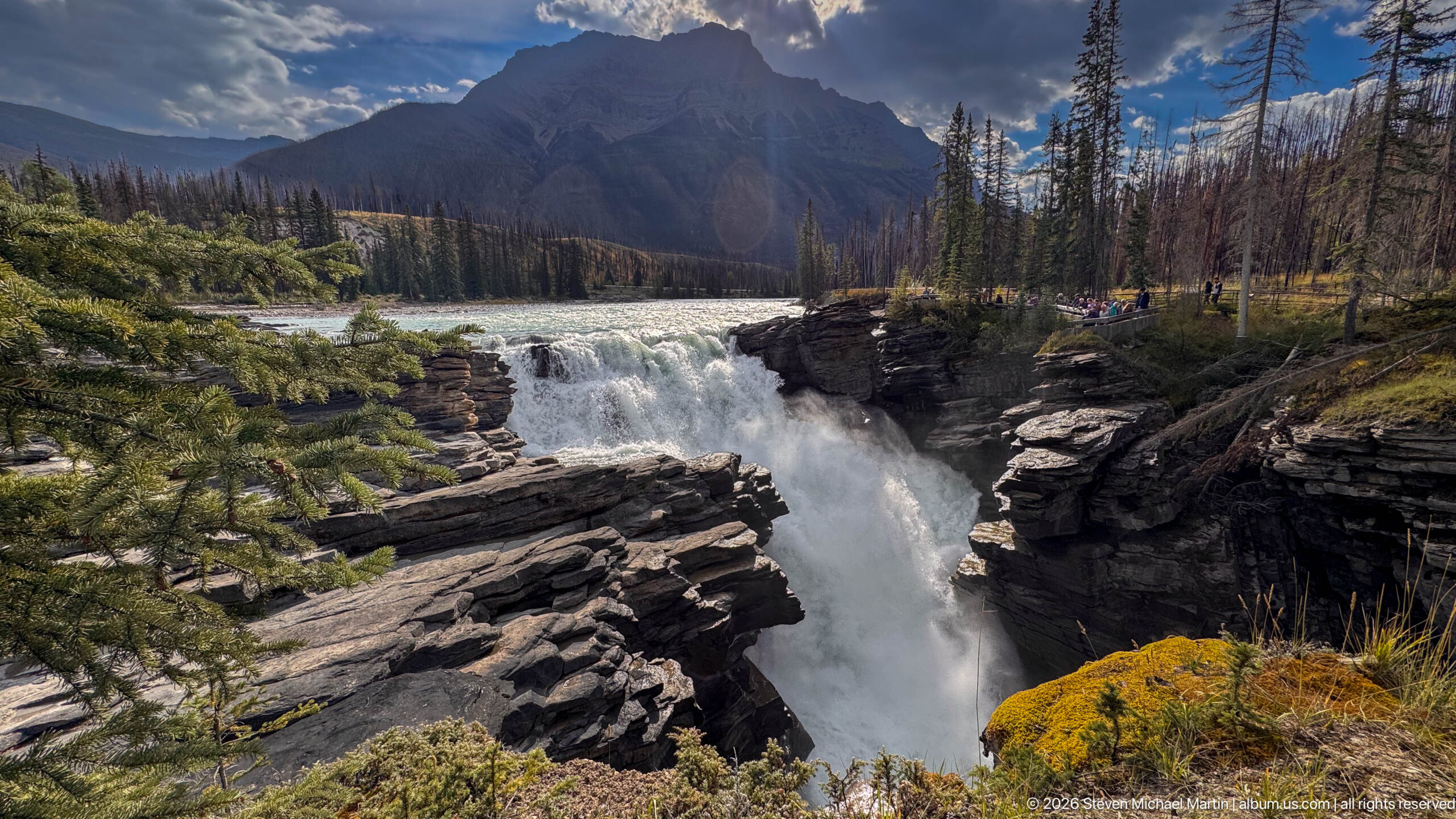 Powerful waterfall plunging into a narrow rocky gorge beneath a mountain, with forest and visitors at right.