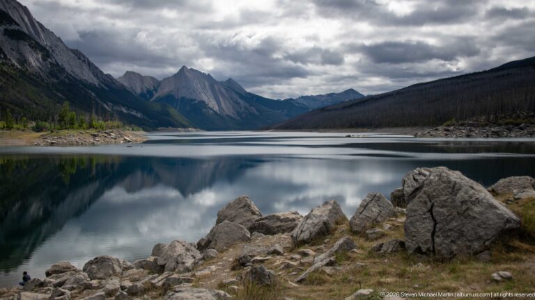Medicine Lake beneath dark storm clouds, with shoreline boulders, mountain walls, and reflections on calm water.