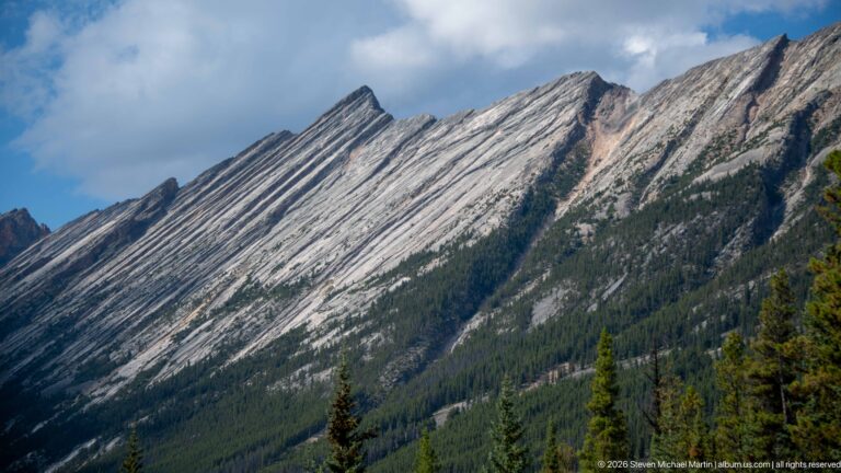 Steep mountain ridge with strongly tilted parallel sedimentary rock layers above dark conifer forest.