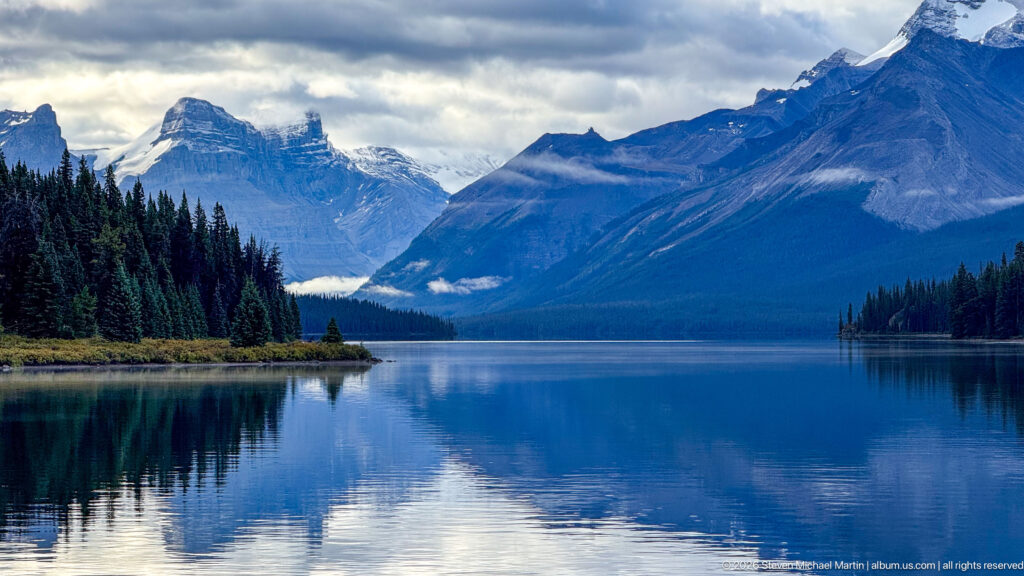 Deep blue Maligne Lake stretches between forested shores toward steep glaciated and snow-streaked mountains beneath low clouds
