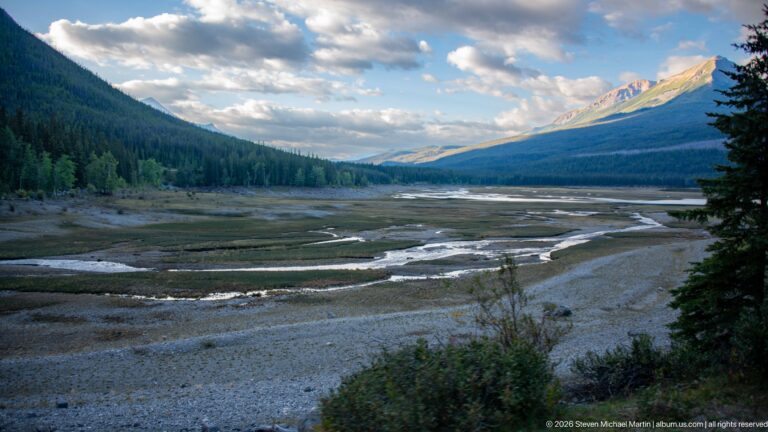 Low-water Medicine Lake basin with exposed gravel flats, shallow channels, forested slopes, and sunlit mountains.