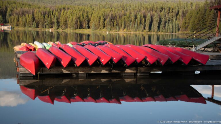 Row of red canoes stored upside down on a rack above calm Maligne Lake with reflections below.