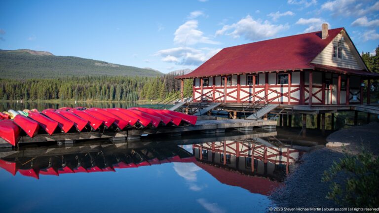 Red canoes on racks beside the Maligne Lake Boat House, with building and boats reflected in calm water.