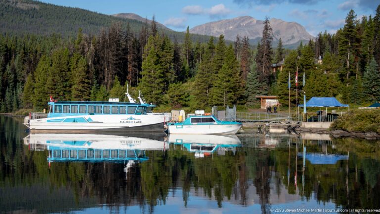 White and blue passenger boats moored at a Maligne Lake dock with forest and mountains behind.