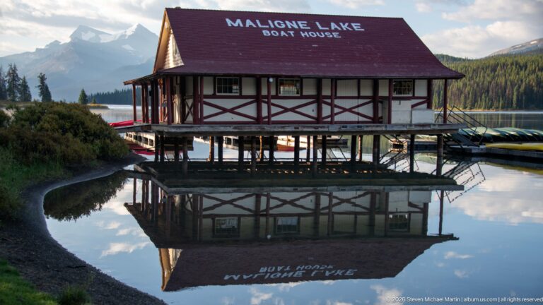 Maligne Lake Boat House with red roof reflected in still water, with canoes stored along the lakeside structure.