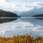 Calm Maligne Lake reflecting clouds, forested shores, and rugged mountains beneath a partly cloudy sky.