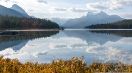 Calm Maligne Lake reflecting clouds, forested shores, and rugged mountains beneath a partly cloudy sky.