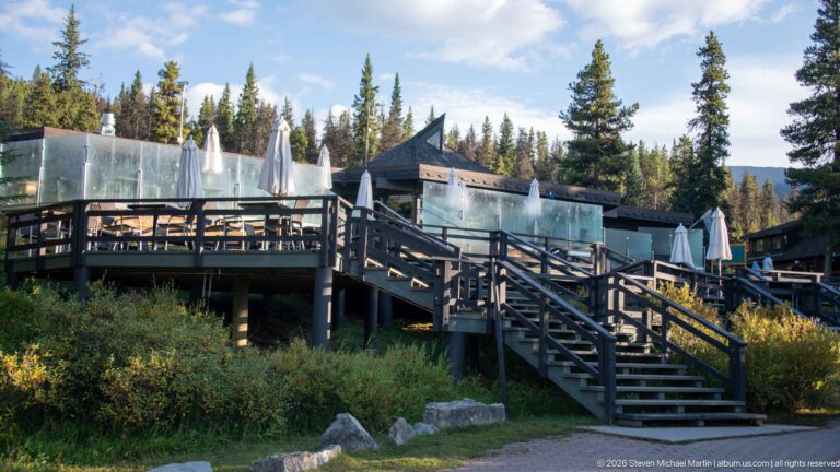 Timber stairs and elevated outdoor terraces with glass screens and umbrellas among conifers at Maligne Lake.