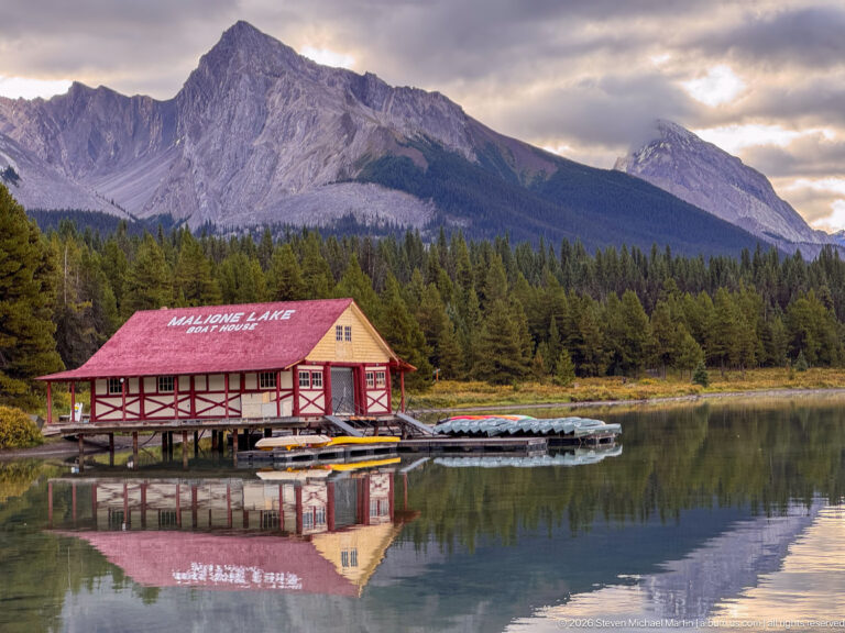 The red-roofed Maligne Lake Boat House and canoe racks reflect in still water beneath a massive gray mountain ridge