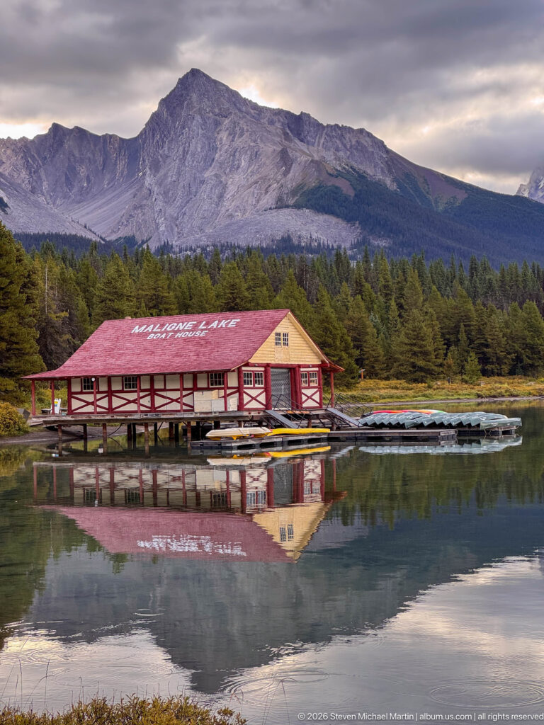 Red-and-cream timber Maligne Lake Boat House stands over calm water with canoe racks, forest, mountain ridge, and a near-complete reflection
