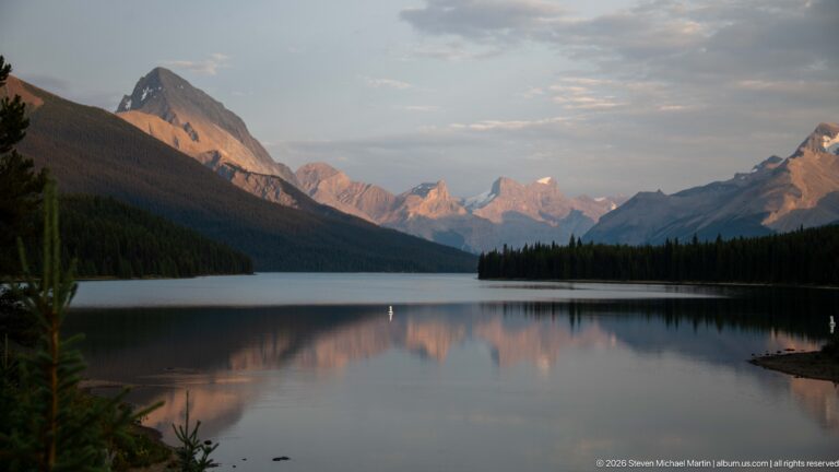 Calm Maligne Lake reflecting forested shores and layered mountains touched by warm evening light.