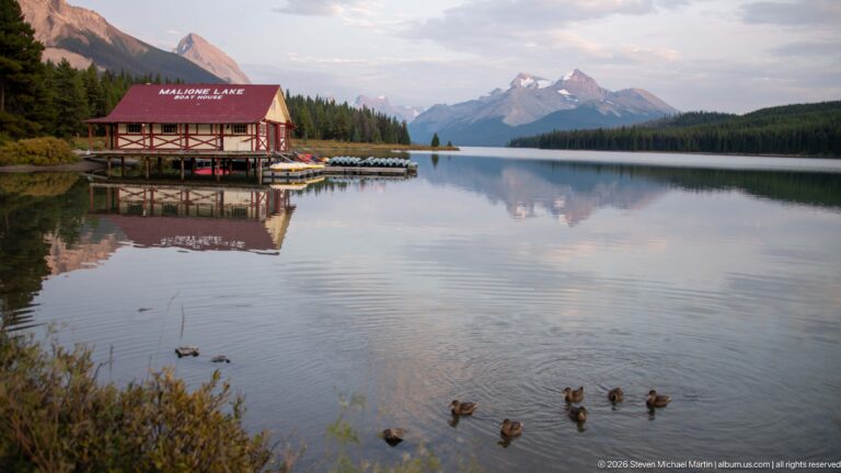 Red-roofed Maligne Lake Boathouse reflected in calm water, with docks, distant mountains, and ducks in the foreground.