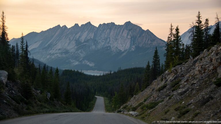 Empty paved road descends through a rocky cut toward conifer forest, distant water, and a jagged mountain ridge.