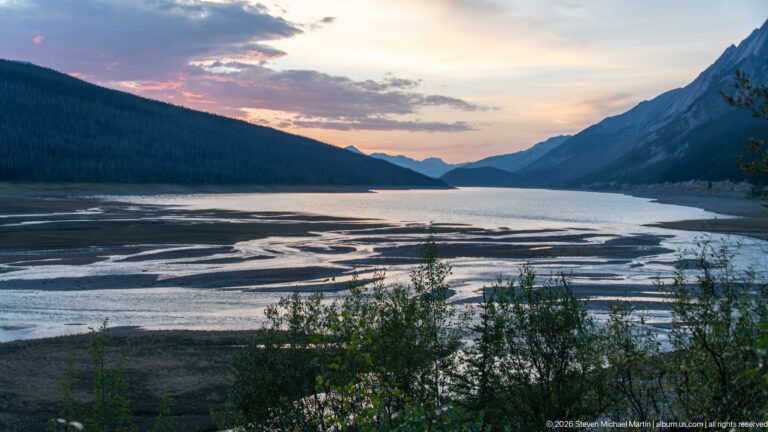 Branching pale channels spread across an exposed mountain lakebed beneath forested slopes and a pastel evening sky.