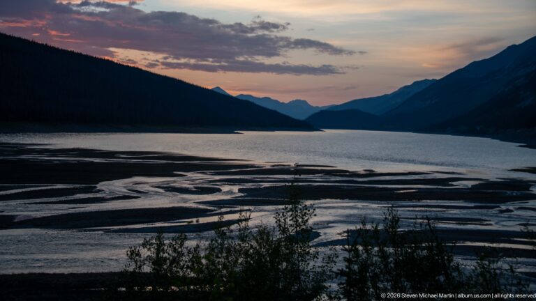 Braided water channels cross an exposed lakebed below dark forested slopes and distant mountains at dusk.