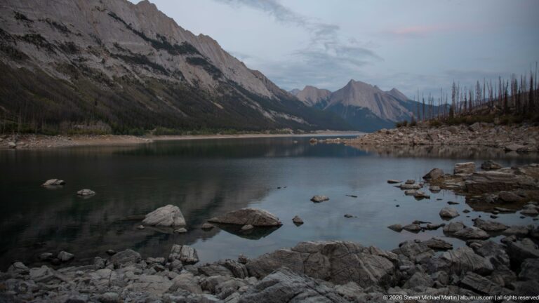Boulders in calm shallow water lead toward an exposed shore and layered mountain peaks in a long valley basin.
