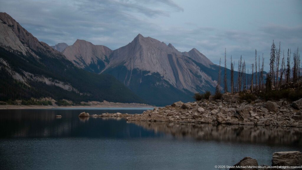 Still mountain water beside a broad rocky shoreline, with steep gray peaks and sparse standing trees beyond.