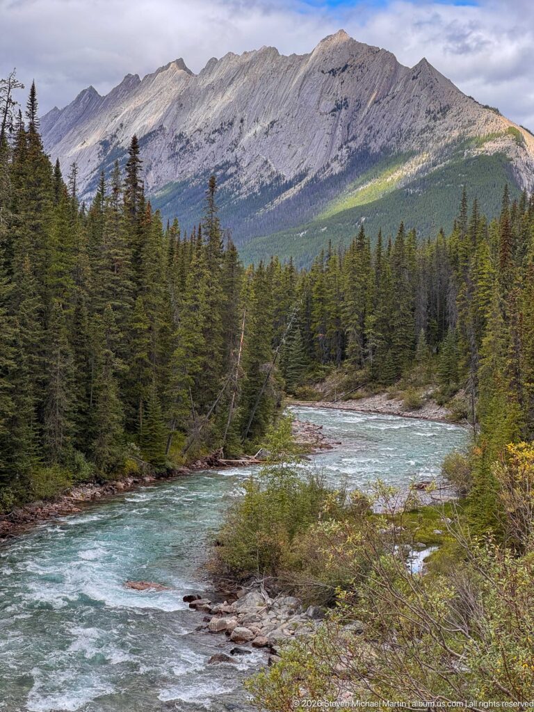 Fast turquoise water bends through dense conifer forest below a broad gray mountain ridge