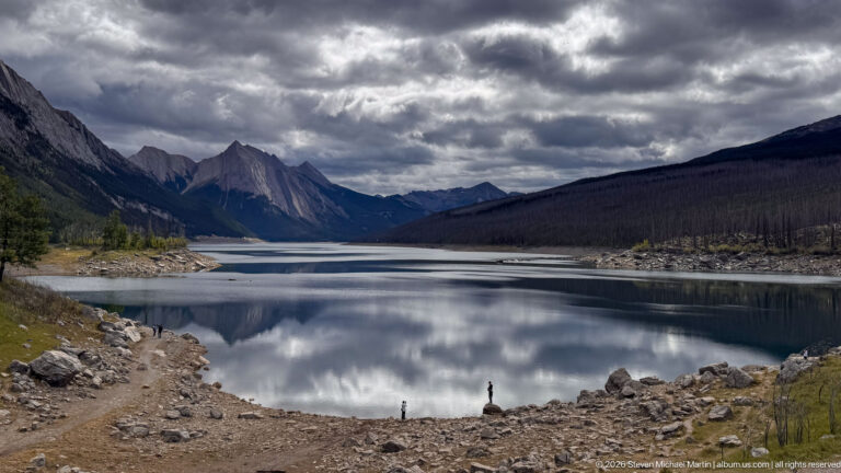 A broad view looks down Medicine Lake toward angular peaks beneath dark clouds, with extensive rocky shoreline exposed around calm reflective water