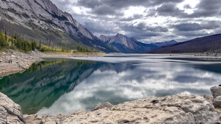 Medicine Lake extends between steep mountain walls beneath heavy clouds, with broad pale exposed shoreline and strong reflections