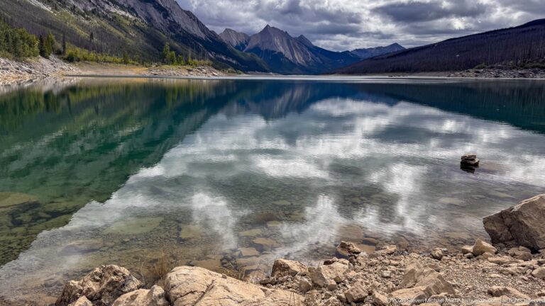 Clear water mirrors dramatic clouds and mountain walls above a rocky foreground shoreline, with low water exposing broad margins around the basin